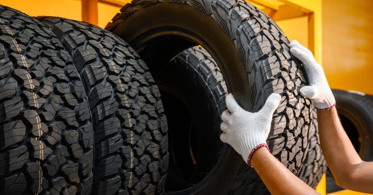 A man wearing white car gloves taking a large, heavy-duty, automotive off-road tire off a yellow storage rack.