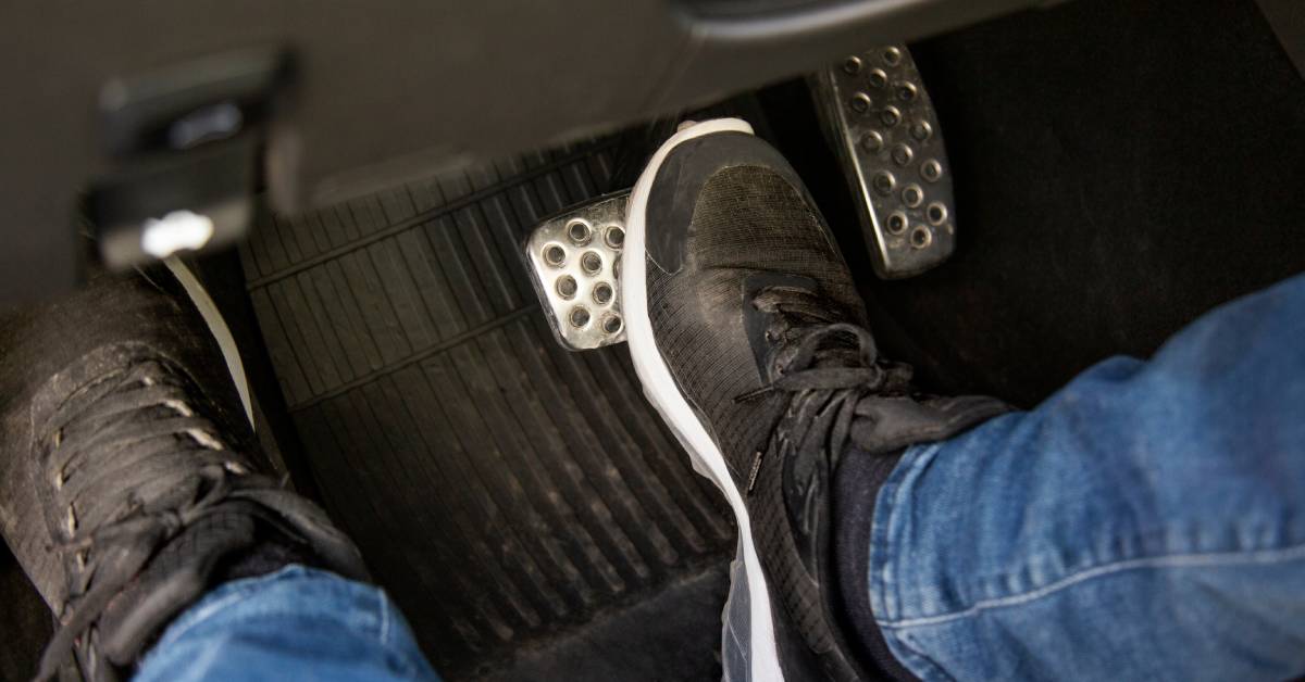 The underfoot compartment of the driver's seat shows two black sneakers engaging the brake and the clutch pedal.