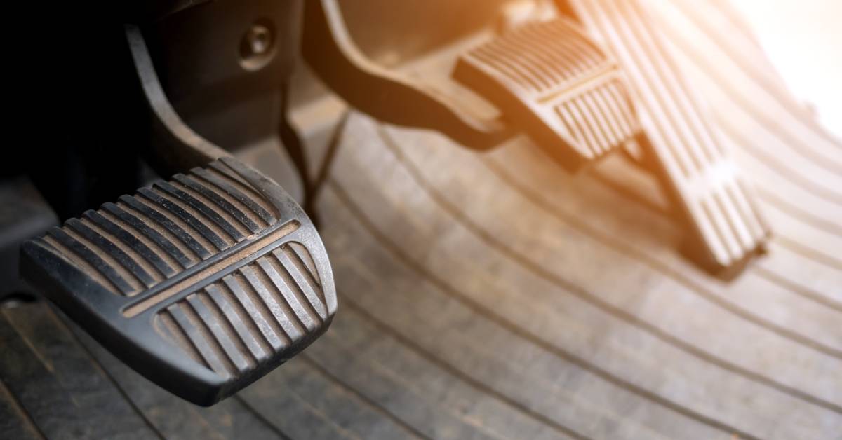 The underfloor of a manual transmission vehicle, featuring the clutch, brake, and gas pedal with a sunset glare.