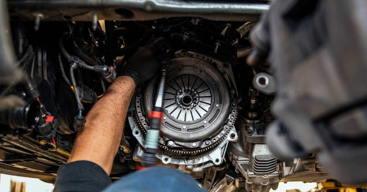 A mechanic in a blue cap holds a tool and stands underneath the clutch disc of an elevated vehicle on a garage lift.