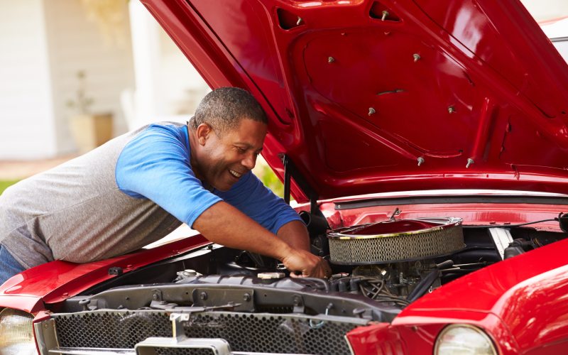 A man leaning over the open engine compartment of a classic red car and reaching within the engine to adjust something.