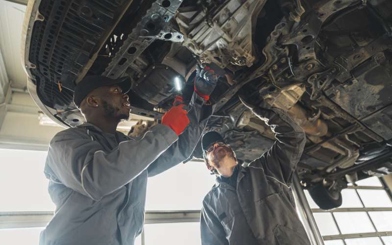 Two auto mechanics inspect the undercarriage and clutch of a car on a lift with their hands and a flashlight.