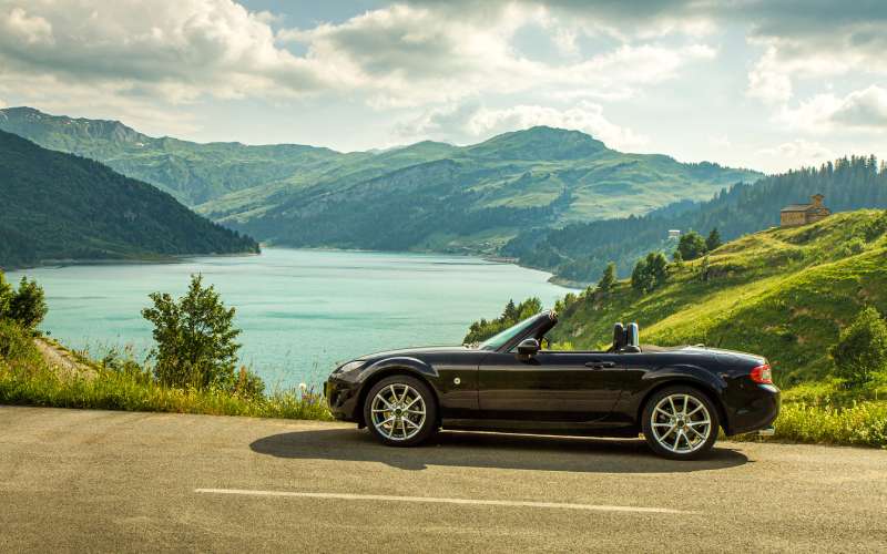 A black Mazda Miata from the side. The top is down and it sits on a road with rolling green hills and a lake behind it.