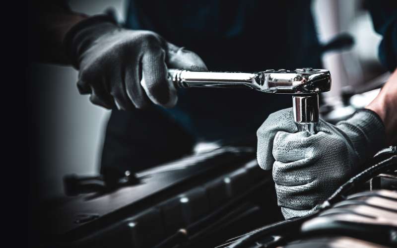 An auto mechanic wearing grey work gloves and using a socket wrench to unscrew something within a car engine.