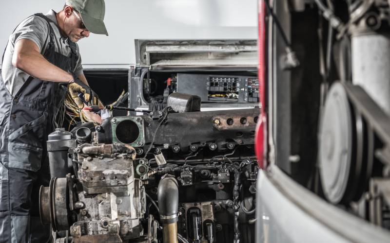 A mechanic wearing overalls, a hat, and gloves works on a disassembled automotive diesel engine in a workshop.