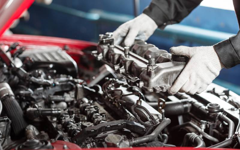 A mechanic wearing gloves holds an engine part over the open engine bay of a red diesel vehicle.