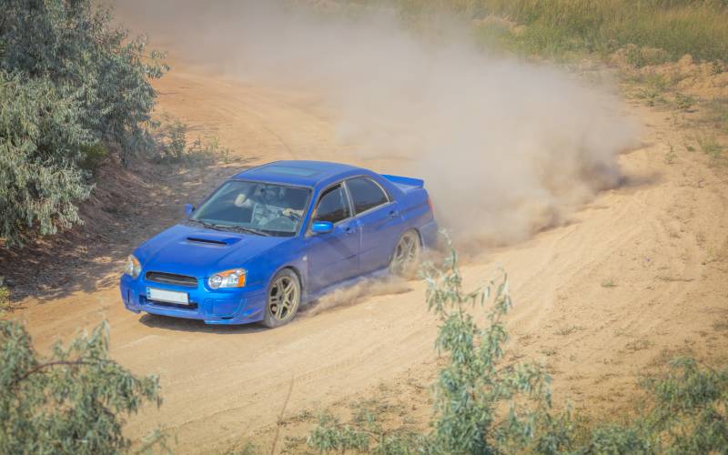 A long-distance shot of a blue Subaru rally car sedan turning on a dirt track corner and kicking dust behind it.