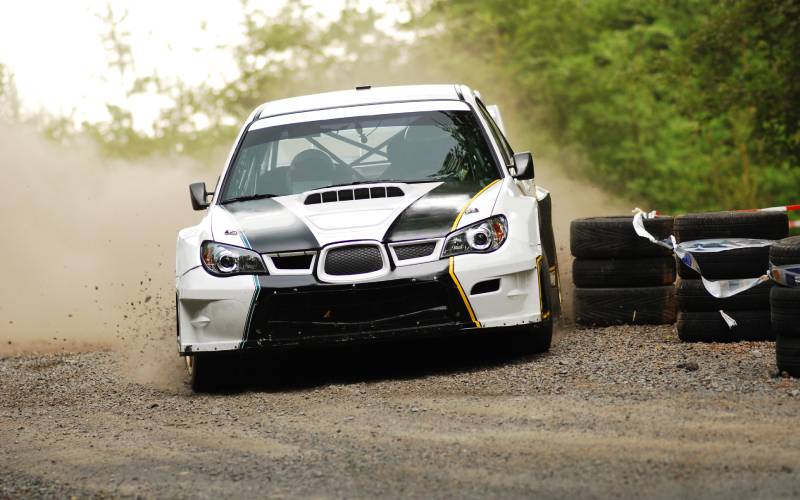 A frontal view of a white and black Subaru World Rally Championship car as it maneuvers around a corner on a dirt track.