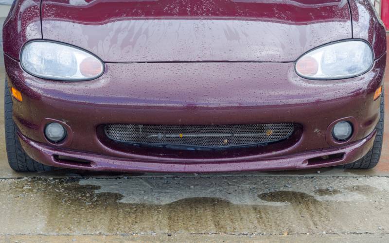 The front bumper, grille, and headlights of a maroon Mazda Miata still dripping after a recent car wash.