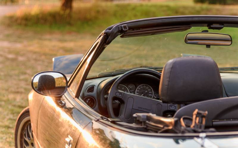 A rear view of a Mazda Miata with the top down and headlights up in a field against the glare of a setting sun.