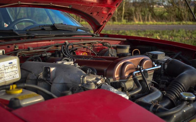 A close-up of a classic red Mazda Miata with its engine bay open showcasing its 1.6-liter engine in a grass field.