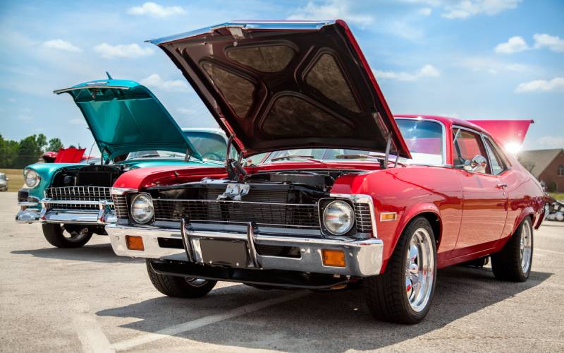 Two vintage muscle cars, one red, and one teal, parked in a parking lot with their hoods open on a sunny day.