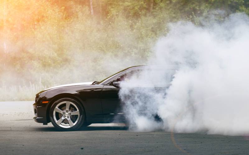 A side profile of a black muscle car spinning its wheels. Smoke obscures the rear end of the vehicle.