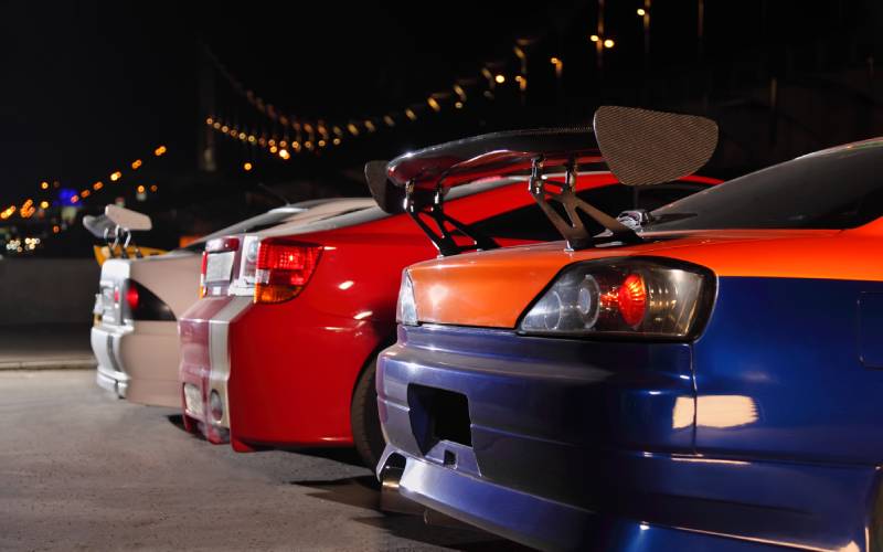 The rear and spoilers of three drift cars parked next to each other at night with an illuminated bridge in the background.