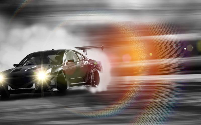 A black sports car drifts around a corner, creating smoke with the vehicle in focus and the surroundings blurred.