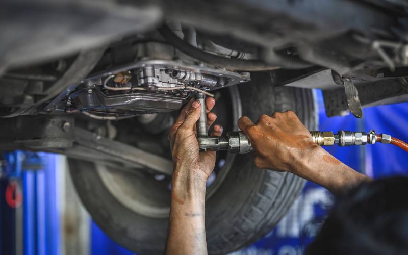 A mechanic's hands reach up to attach a pressurized tool to a part underneath a lifted car in a service garage.