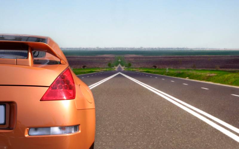 The backside of an orange sports car with a spoiler driving down an empty road. The street is surrounded by fields.