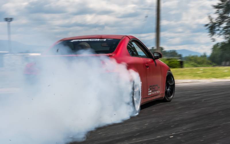 A rear view of a red sports car drifting around a corner of a race track and creating white smoke from the wheels.