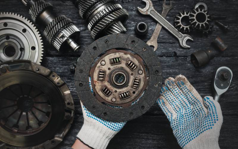 An overhead view of an old and used car clutch disc held by gloved hands over a table of car clutch components.