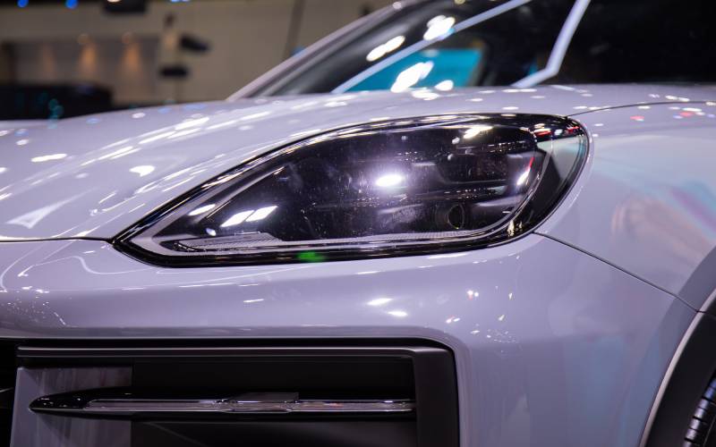 A close-up view of the headlight of a white Porsche car in a showroom with interior lights reflected off the headlight.