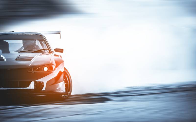 A sports car drifting on a race track and producing lots of smoke with a helmeted driver behind the wheel.