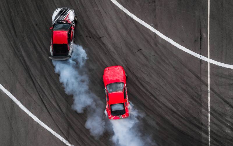 An aerial view of two race cars racing around a track. Both cars are turning a corner and producing smoke.