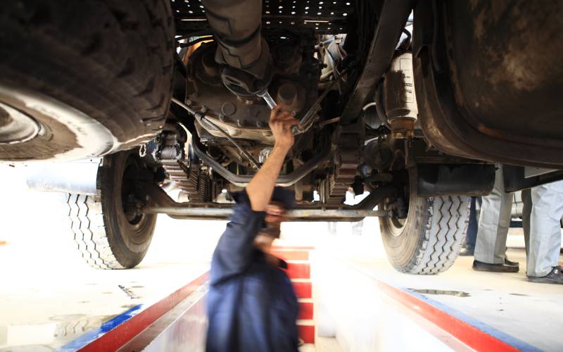 An automotive mechanic in a blue work suit stands underneath a pickup truck chassis and adjusts something with a wrench.