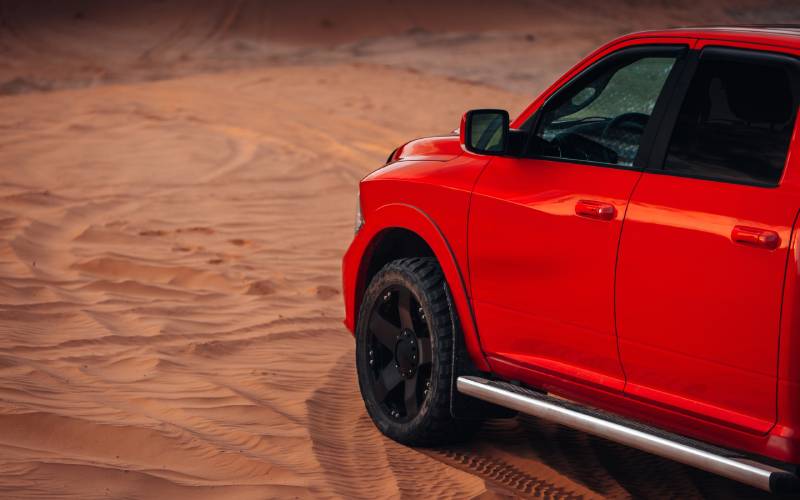 A red heavy-duty four-door pickup truck with black rims and running board faces away on a sandy dune.