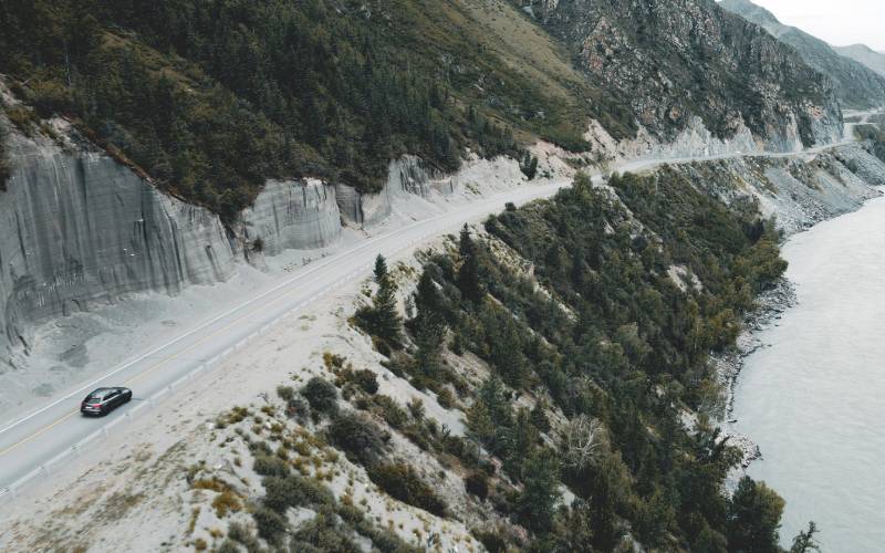 A wide landscape view of a grey Audi car driving on a two-lane road against a cliffside overlooking water.