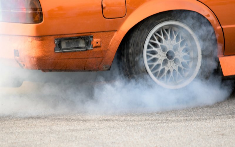 A close-up of the rear wheel and tire of an orange car spinning its wheels on the pavement and generating smoke.