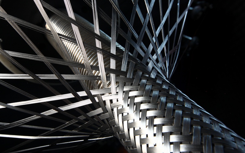 A close-up of a grooved cylinder being wrapped in stainless steel braiding against a black background.