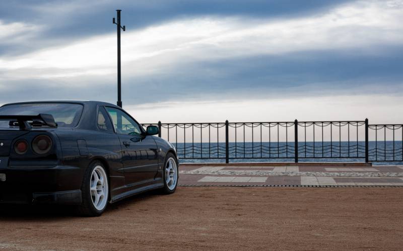 A rear view of a black Nissan Skyline with a spoiler parked on a dirt lot overlooking the ocean on a cloudy day.