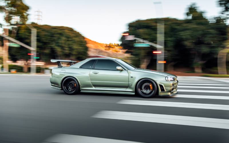 A side-view of an in-focus metallic green Nissan Skyline with a spoiler driving through an intersection.