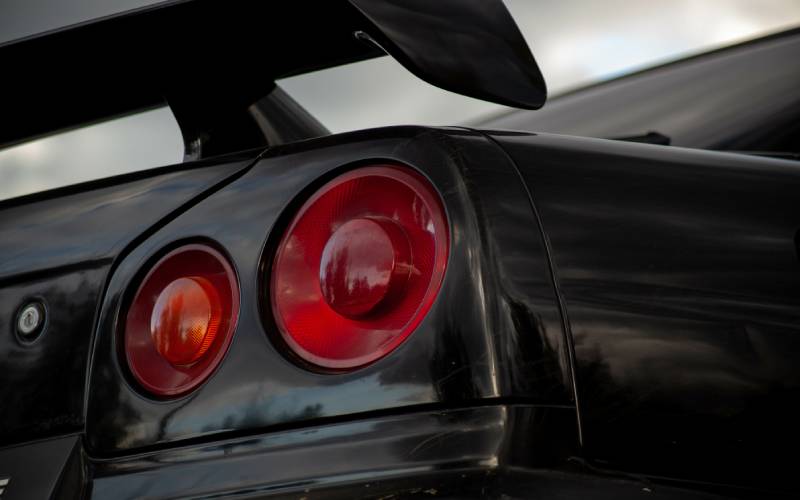 A close-up of the rear taillights of a black Nissan Skyline with a spoiler. The taillights feature two circular red lights.