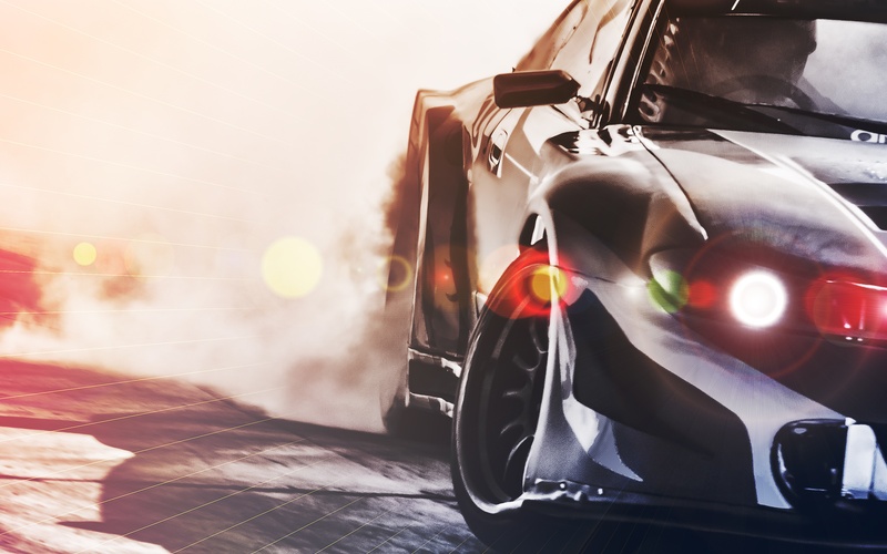 A close-up of a black sports car drifting around a corner with smoke coming from the rear wheels and headlights activated.