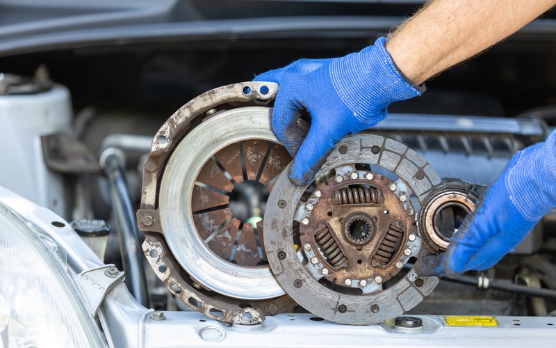 A close-up of a person's hands in blue gloves holding up a used clutch disc, pressure plate, and release bearing of a vehicle.