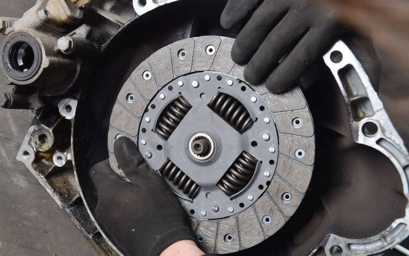 A close-up of a pair of hands with black gloves removing the clutch plate and disc from a vehicle's transmission.