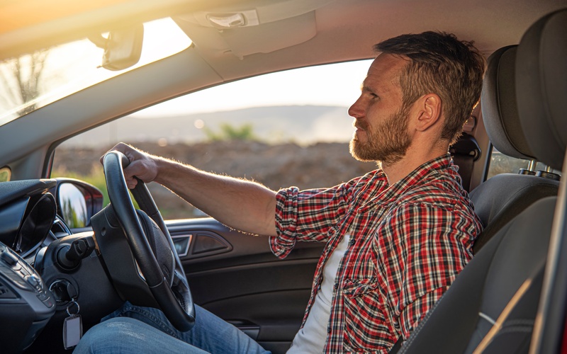 A side view from the passenger seat of a man in a flannel shirt and jeans driving a car while the sun sets.