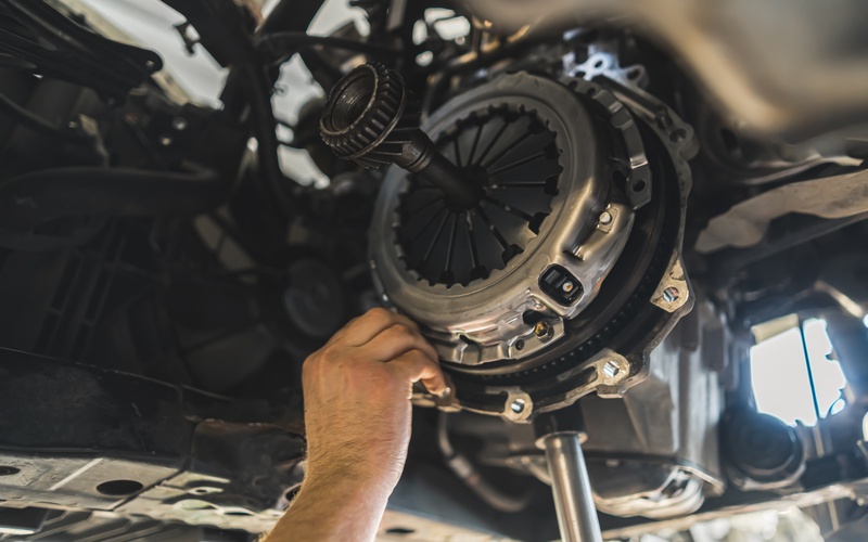 A close-up of a mechanic's hand reaching into the undercarriage of a vehicle on a lift to handle the automotive clutch.