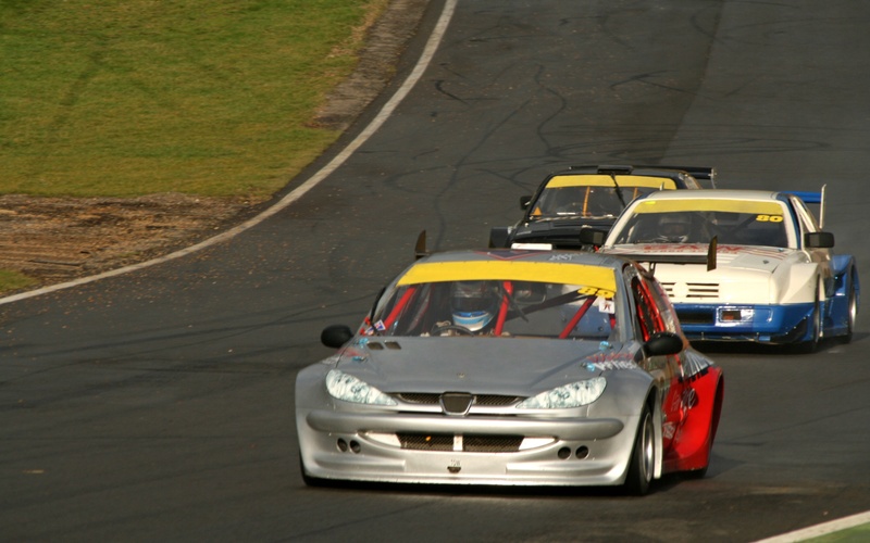 Three spec-production race cars competing on a track. The lead car is silver, the others white and black.