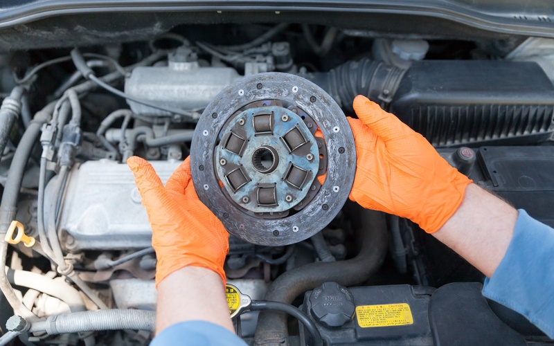 A mechanic's hands with orange latex gloves holding up a used clutch disc friction plate over an open car engine compartment.