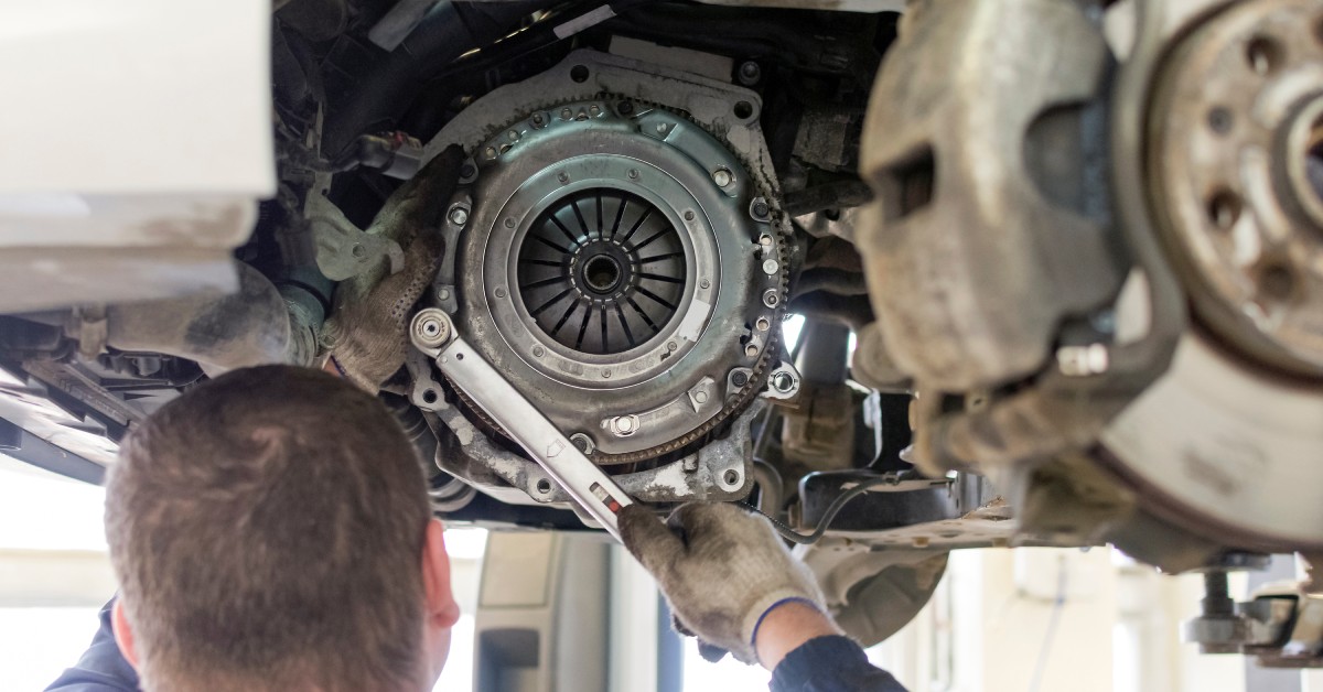 A mechanic with gloves and socket wrench working on the gearbox and clutch of a car elevated on a lift.