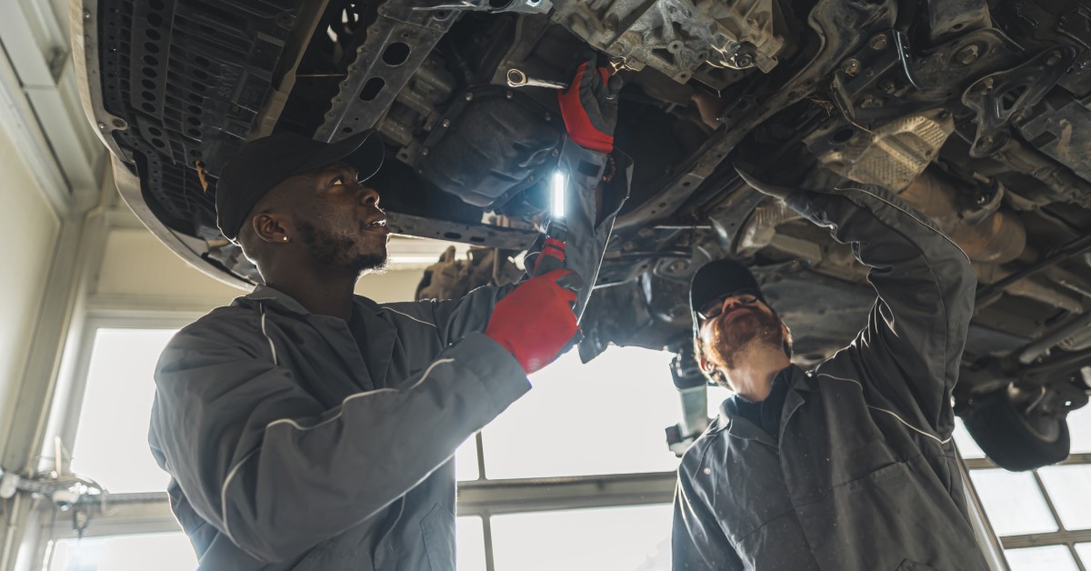 Two mechanics, one holding a flashlight, work on replacing the clutch of a car elevated on a lift in an auto repair shop.