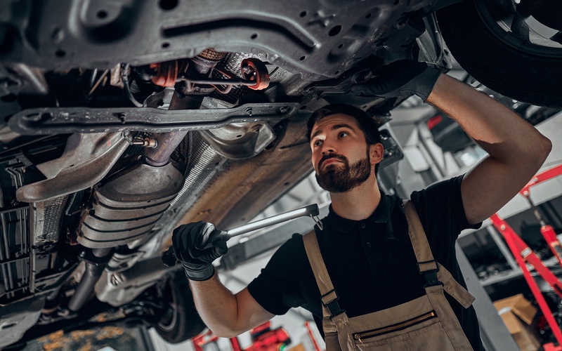 A male mechanic examines the undercarriage of a car on a hydraulic lift in a repair garage with a socket wrench in hand.