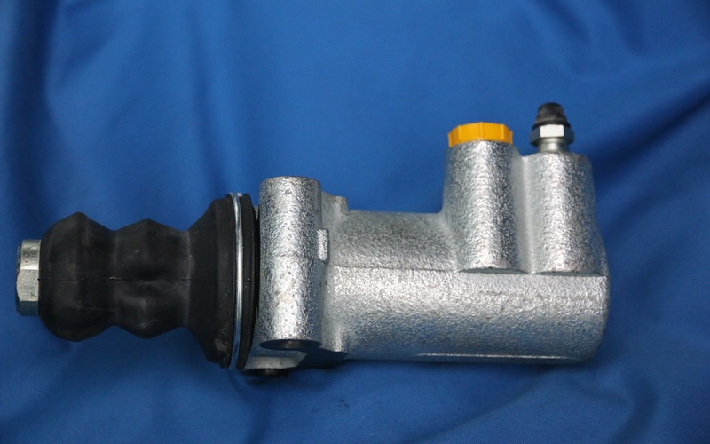 A close-up of a vehicle's new clutch slave cylinder with an iron frame and yellow cap against a blue background.