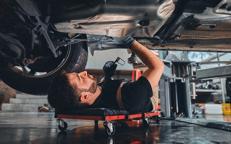 A car mechanic on a creeper examining the undercarriage of a car in a mechanic shop with a wrench tool in one hand.