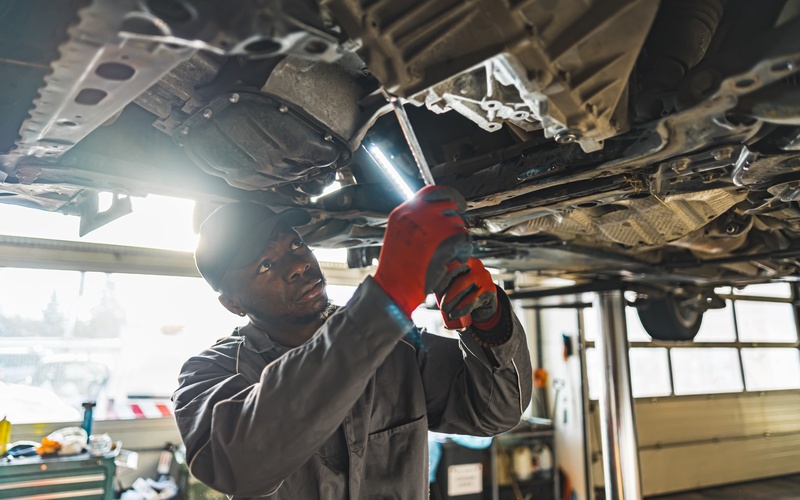 A young auto mechanic in a work shirt, red gloves, and a black hat uses a wrench to work on the undercarriage of a car.