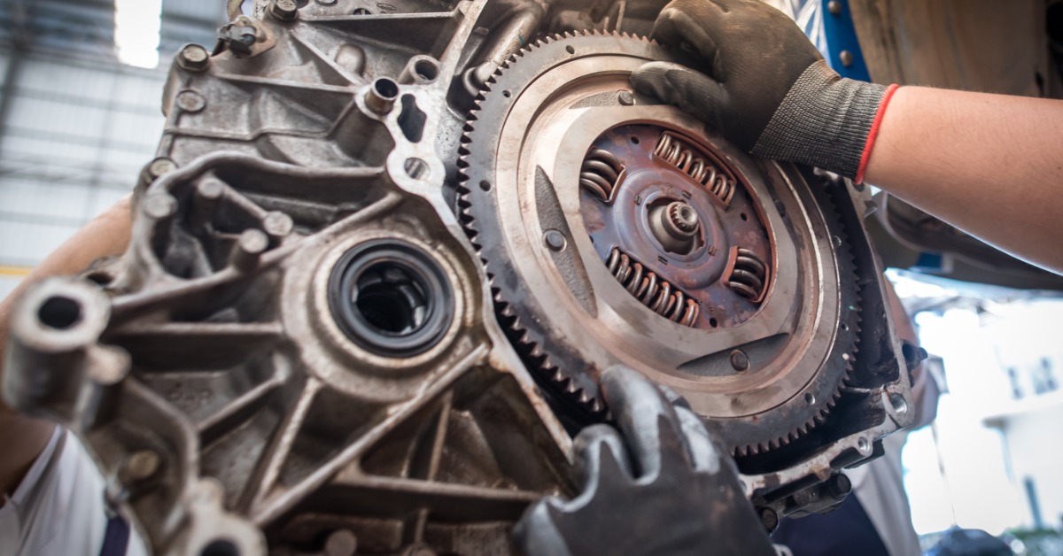 A close-up of a pair of hands handling a car clutch and flywheel removed from a vehicle in a garage.