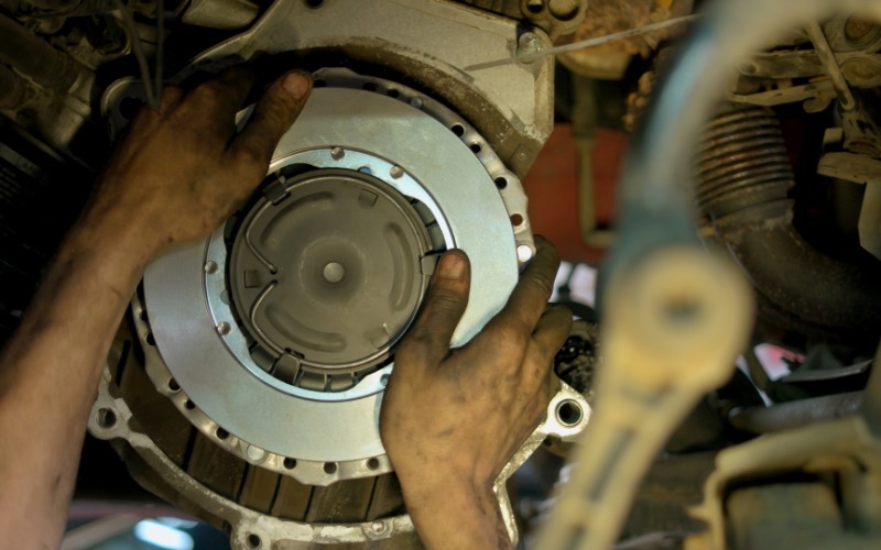 A close-up of the hands of a person reaching into the undercarriage of a car, elevated on a lift, to grab the clutch disc.