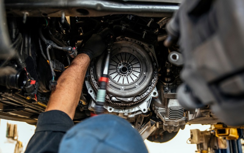 Looking over the head of a mechanic in a blue hat, adjusting the clutch of a car elevated on a lift with a tool.